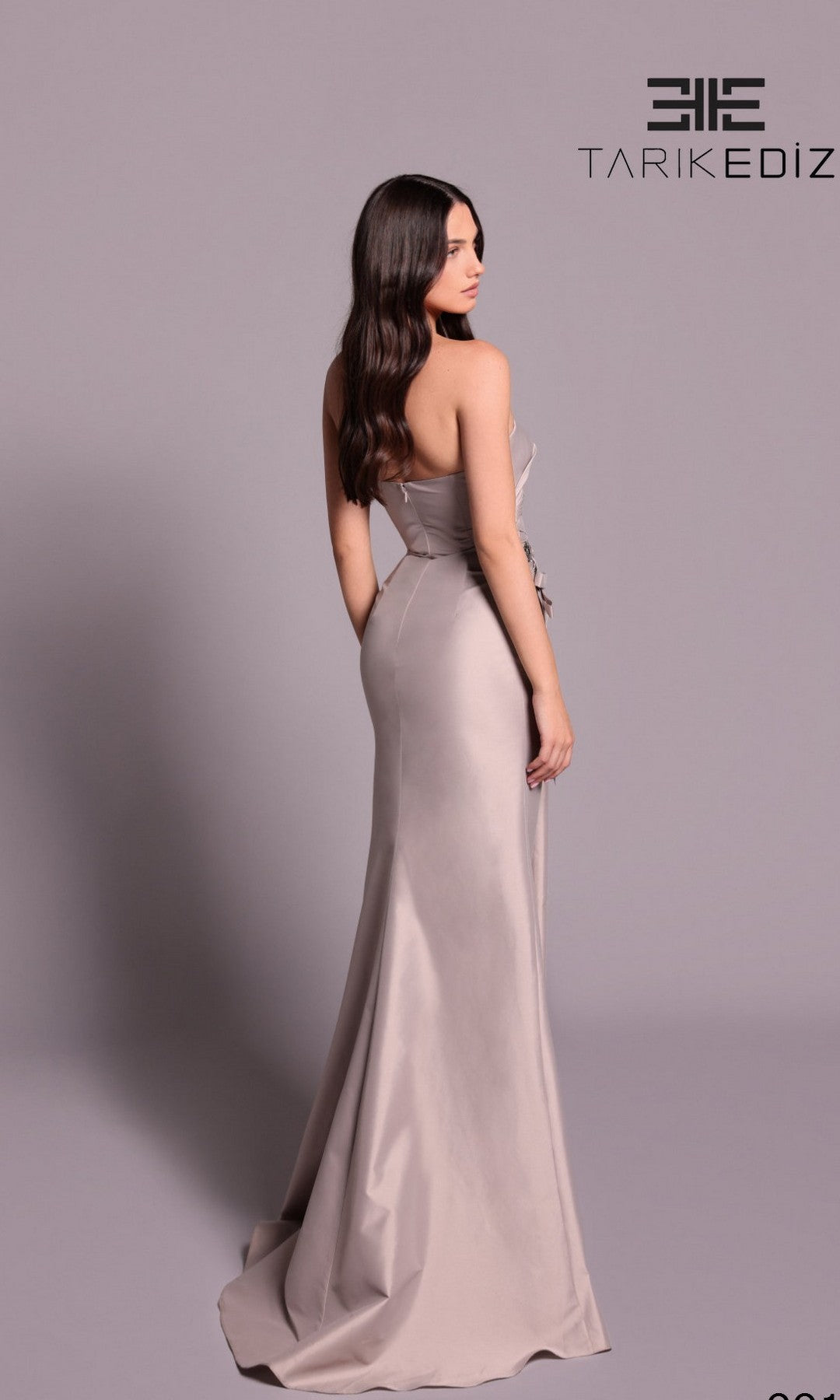 A woman in the Tarik Ediz 99126 long formal dress, strapless and fitted, stands facing away against a plain background, highlighting the gown's elegant floor-length design with the Tarik Ediz logo at the top right.